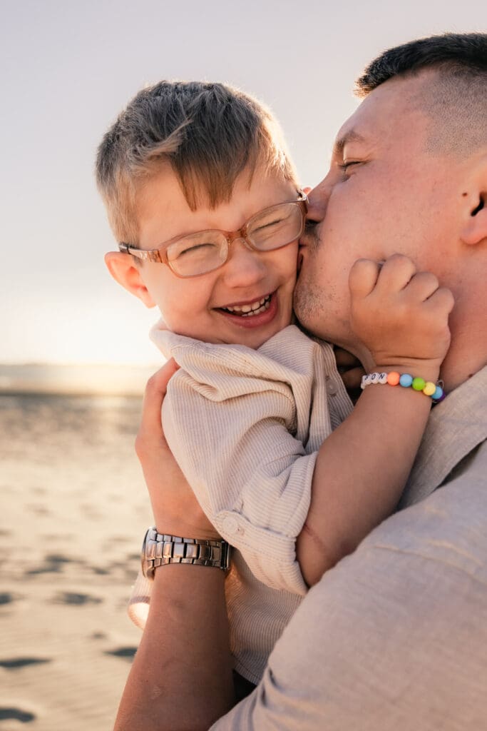 Dad giving boy big kiss on cheek while he laughs at sunset west witterings beach, having fun family photos.