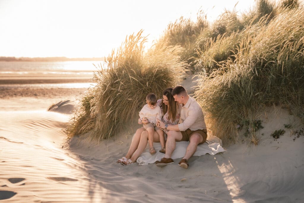 Family of three sitting amongst the sand dunes on west witterings beach in west sussex, with the sun sparkling and setting on the sea behind them