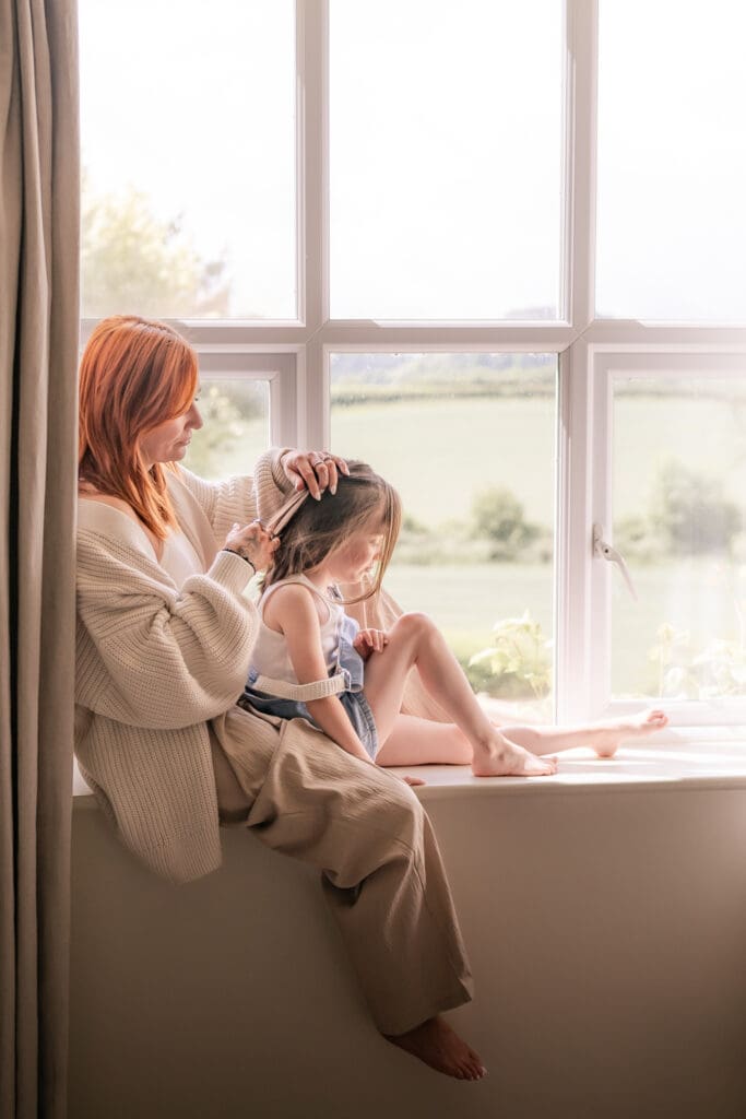Mum gently braiding daughter’s hair by window during Sussex family shoot at home