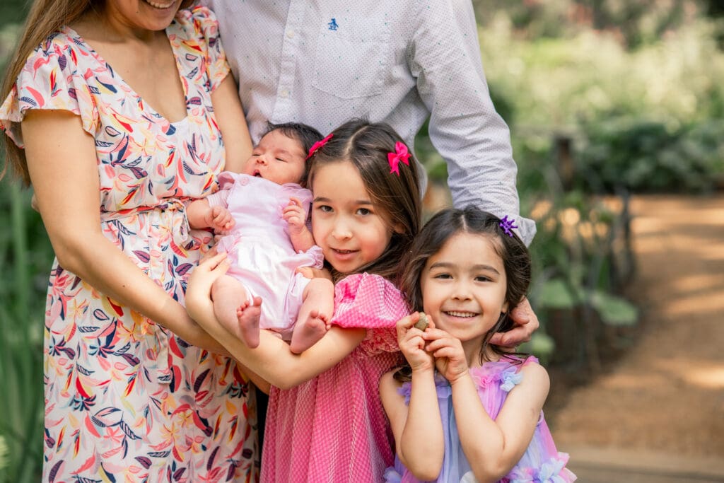 Family with three young daughters during outdoor newborn photoshoot in Sussex, parents holding newborn while siblings smile at camera.