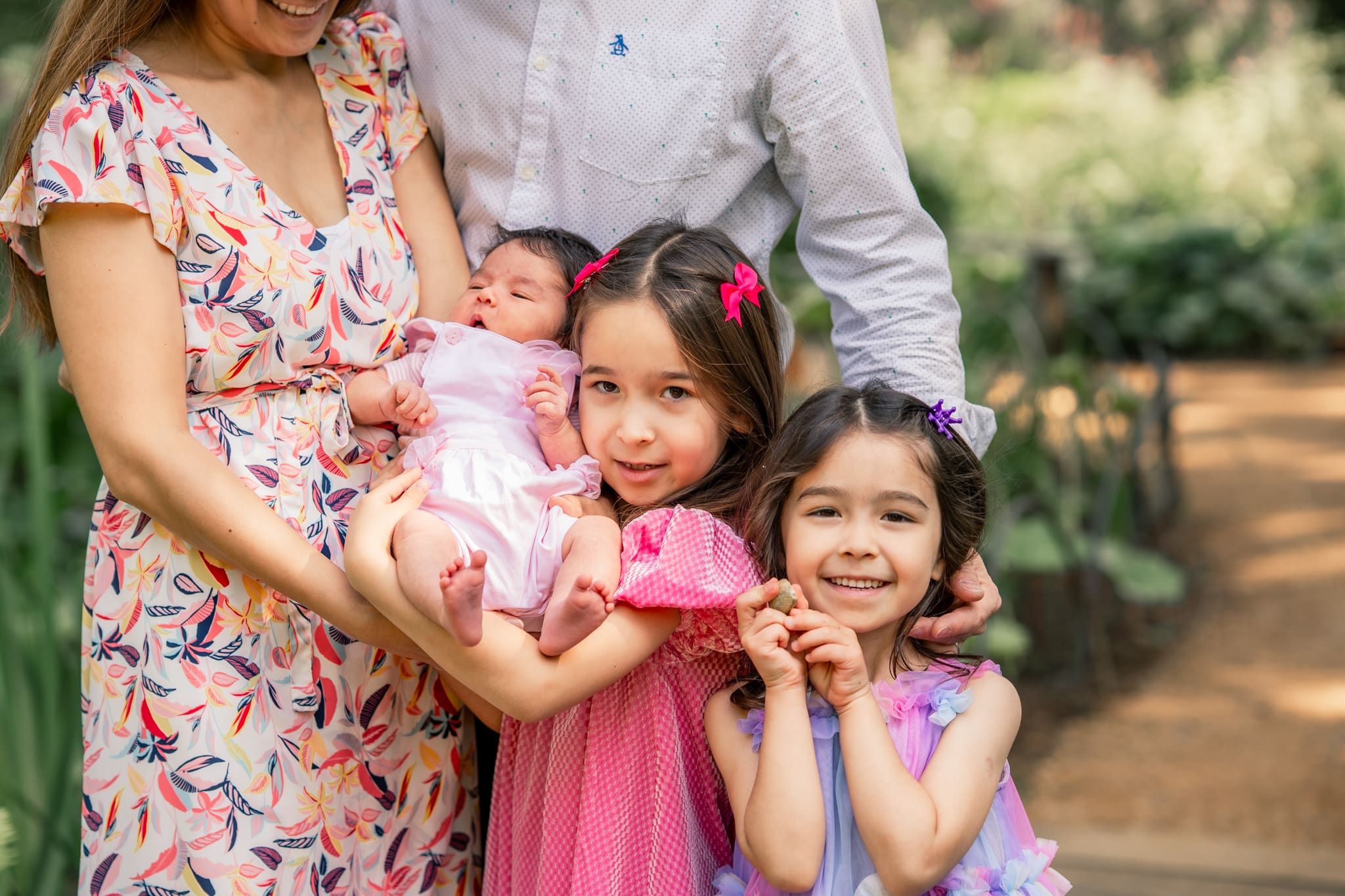 Family with three young daughters during outdoor newborn photoshoot in Sussex, parents holding newborn while siblings smile at camera.