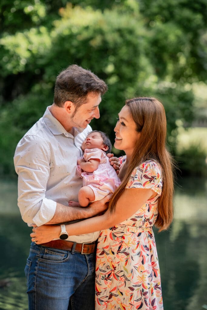 Parents smiling at each other while holding newborn baby during outdoor family session in Sussex.