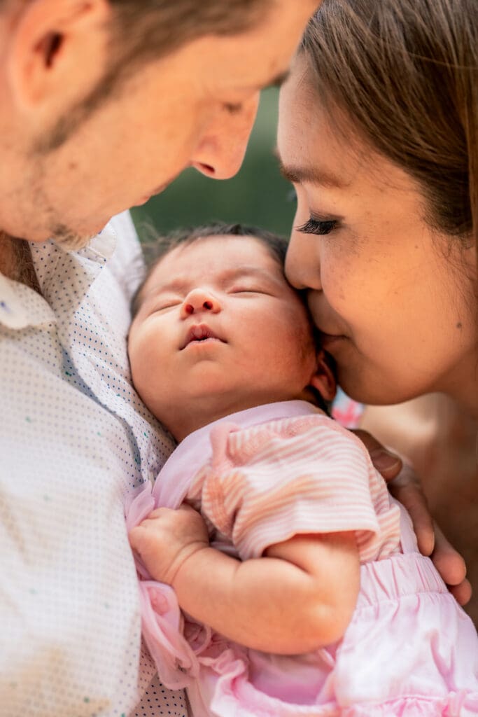 Close-up of parents leaning in to kiss their newborn during outdoor newborn photoshoot in Sussex.