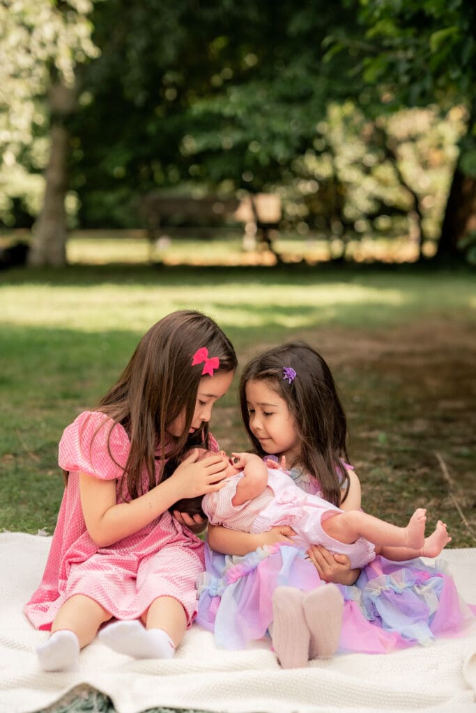 Two young sisters sitting on blanket outdoors, holding and admiring their newborn baby sister during newborn photoshoot in Sussex.