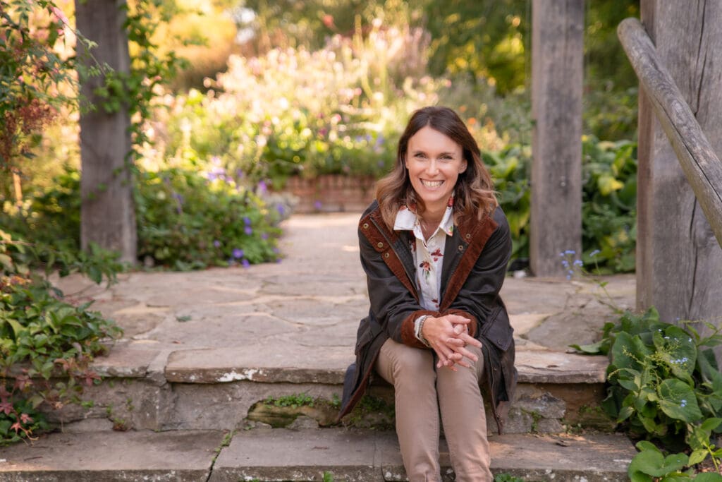 Outdoor branding image of a woman in a peaceful garden setting, dressed casually and seated on steps.