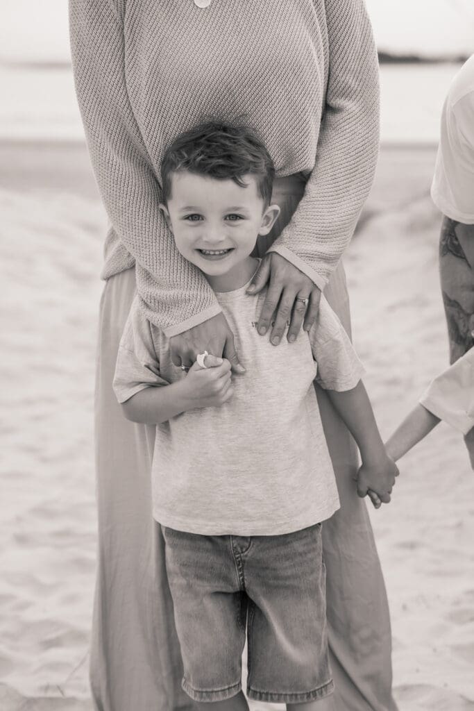 Black and white image of a boy smiling as an adult hugs him from behind at West Wittering Beach.