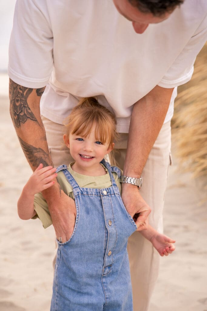 Smiling girl in denim overalls held by her dad during a relaxed family photoshoot on a West Sussex beach.