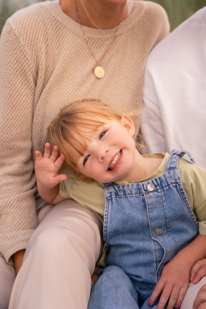 Smiling child waving while sitting on an adult’s lap during a relaxed West Sussex beach photoshoot.