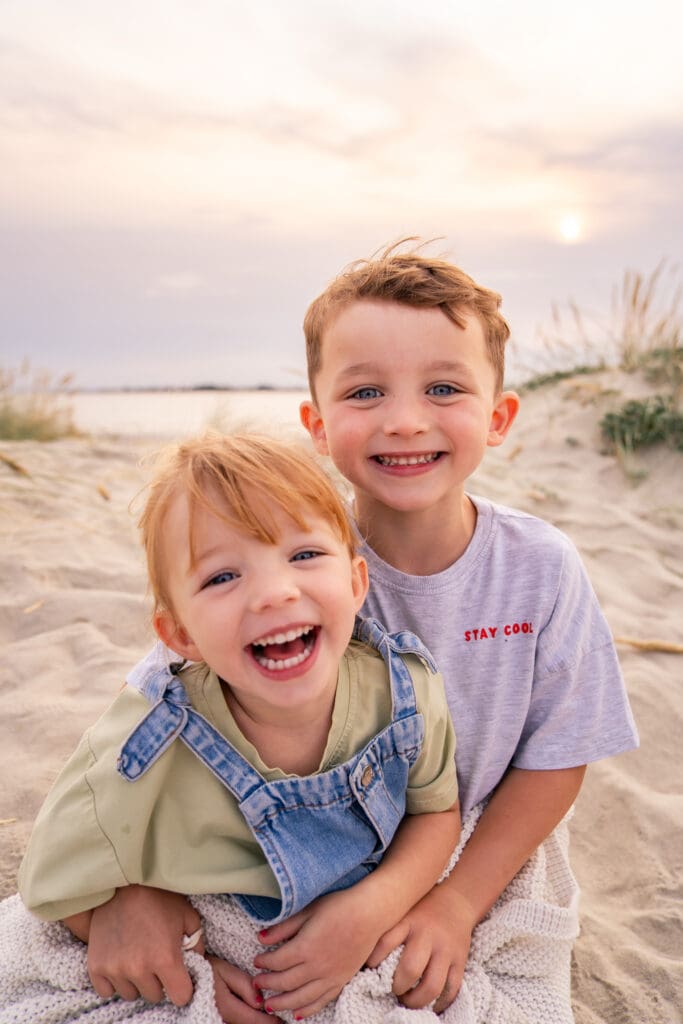 Two siblings smiling and hugging on the sand during a West Sussex family photography session.