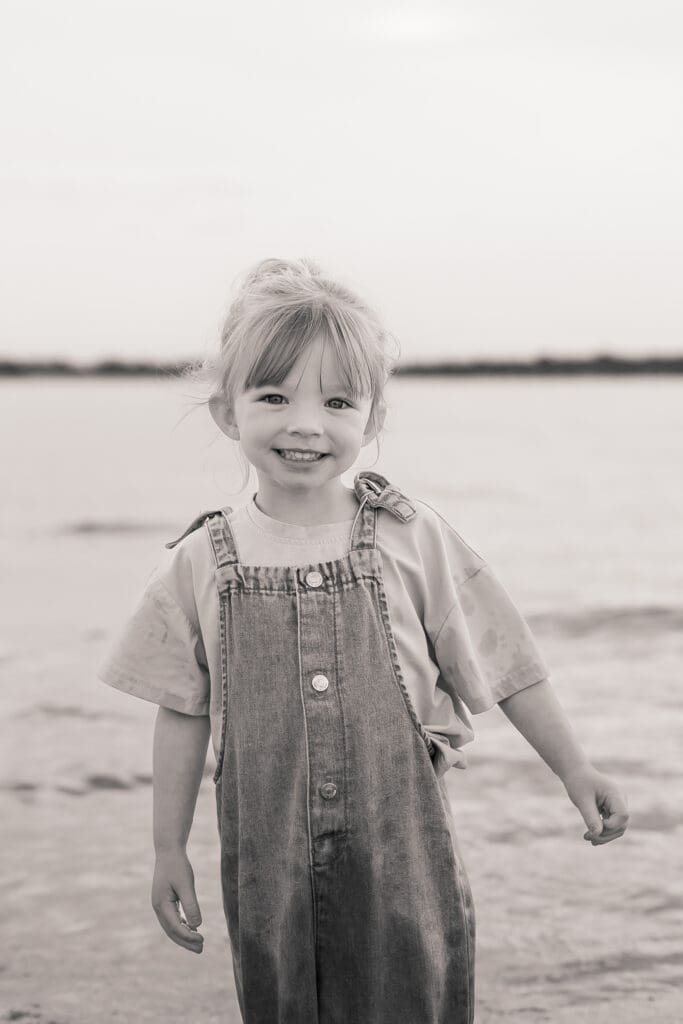 Black and white photo of a child smiling while standing on wet sand at West Wittering Beach.