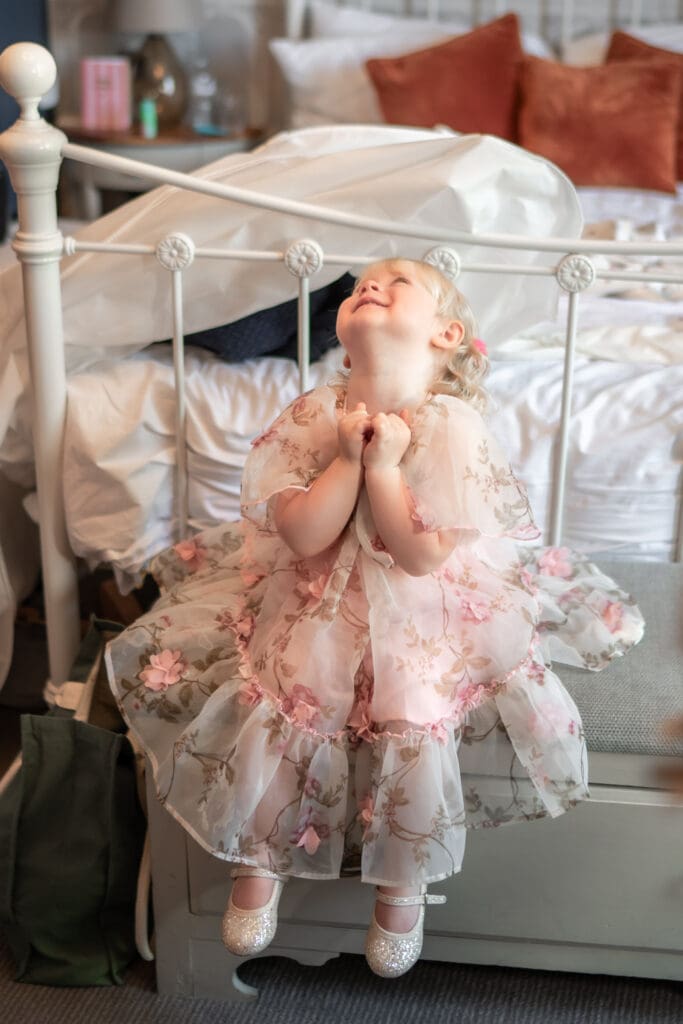 Flower girl in floral dress sitting on bed laughing during wedding morning preparations | Sussex wedding photographer