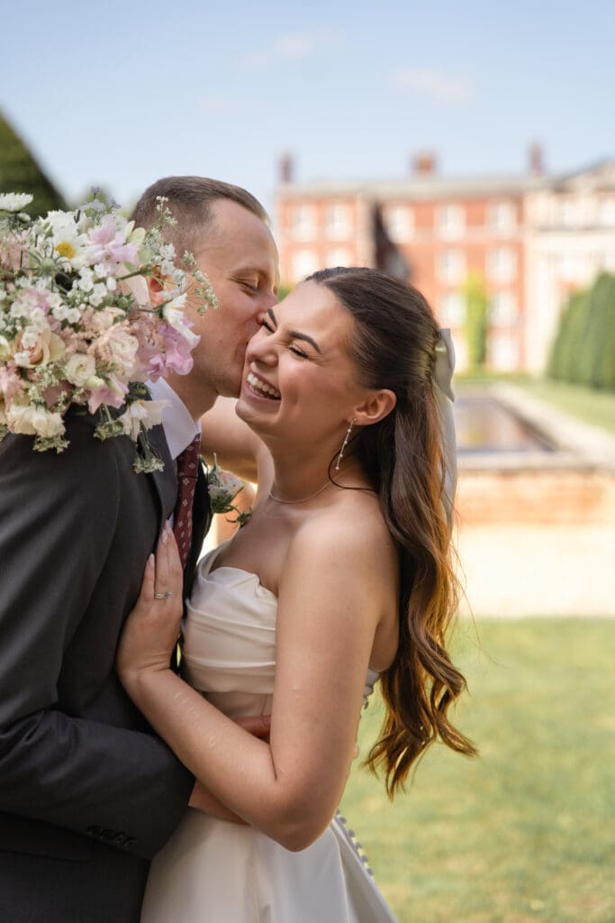 Groom kissing bride's cheek as she laughs during wedding portraits at country house | natural wedding photographer Sussex