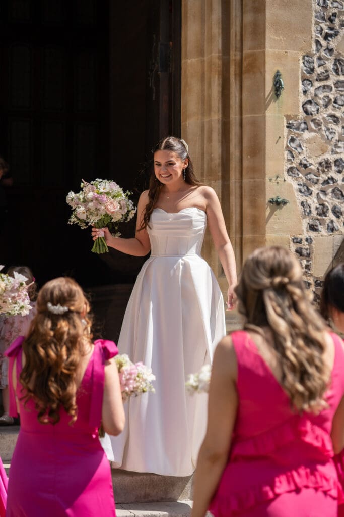 Bride smiling on church steps with bridesmaids in pink dresses | natural wedding photography Sussex