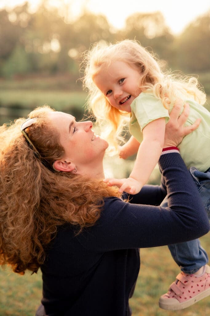 A mother with curly red hair lifting her smiling blonde toddler into the air, both laughing, golden evening light behind them