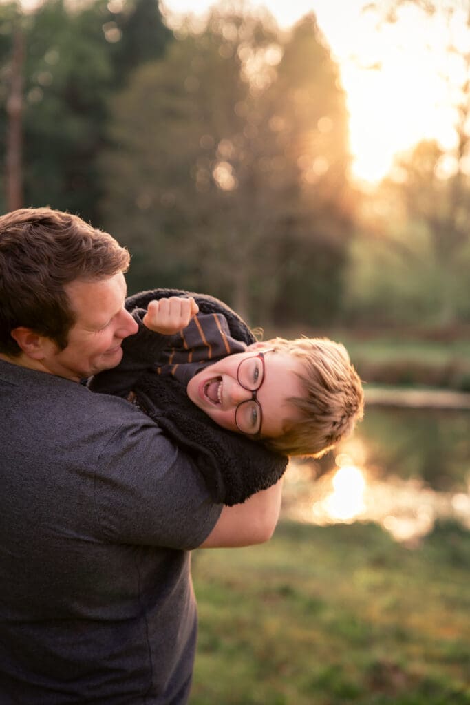 A father bear-hugging his young son in glasses who is laughing with his head thrown back, warm sunset light over a lake