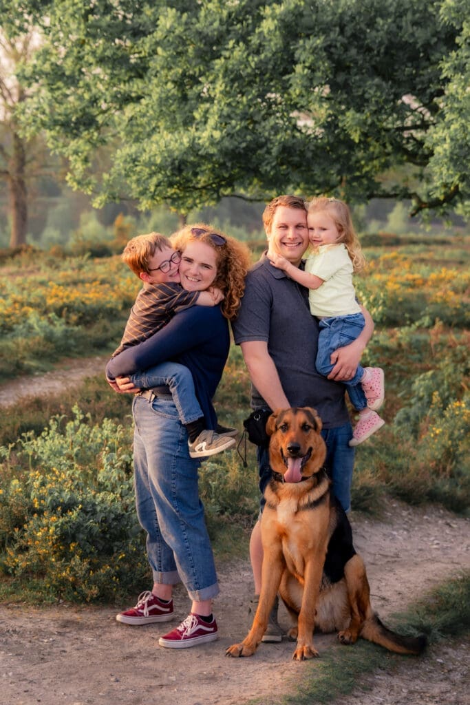A family of four - mum, dad, two young children - smiling and cuddling together with their German Shepherd dog among yellow gorse at sunset