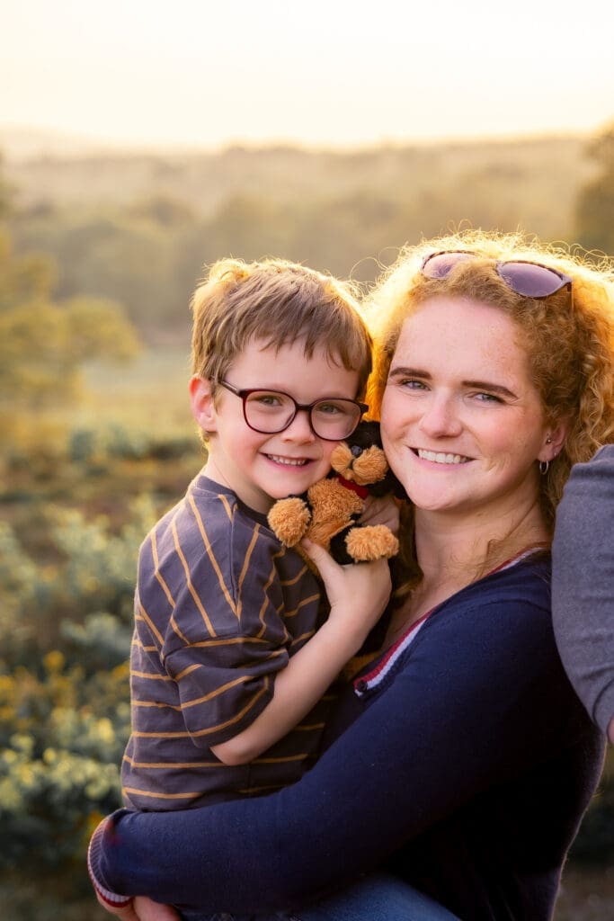 A young boy in glasses clutching a toy bear, cheek to cheek with his smiling mum, South Downs heathland glowing behind them