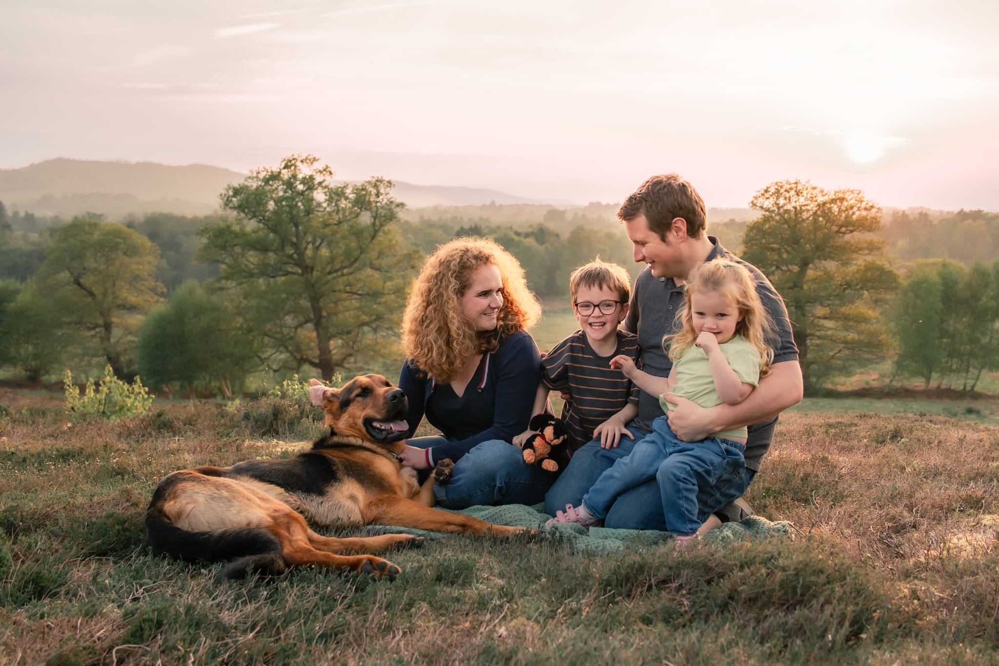 A family of four sitting together on a blanket at sunset, laughing as their German Shepherd dog rolls over demanding attention, rolling hills in the background