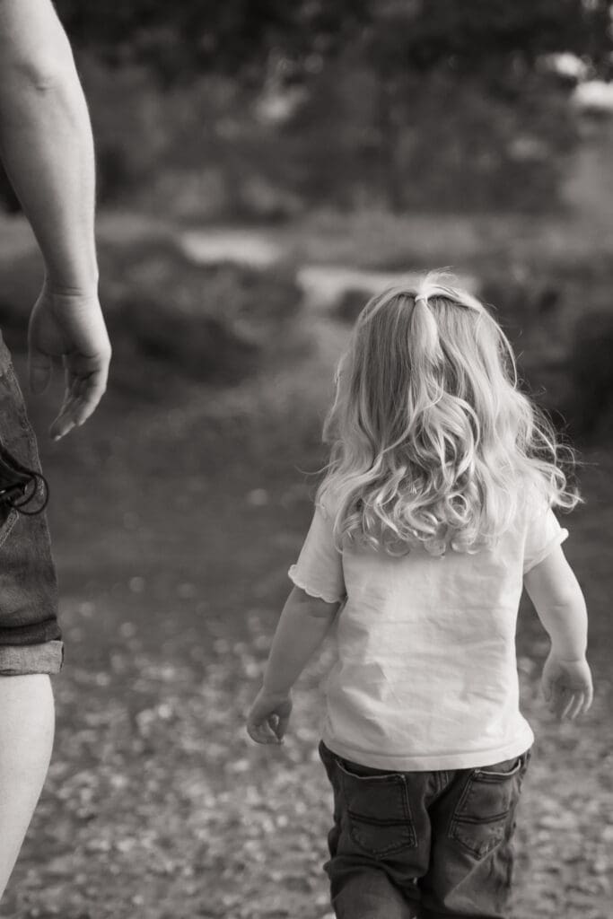 Black and white image of a toddler girl with curly hair walking away along a path, a parent's leg visible beside her