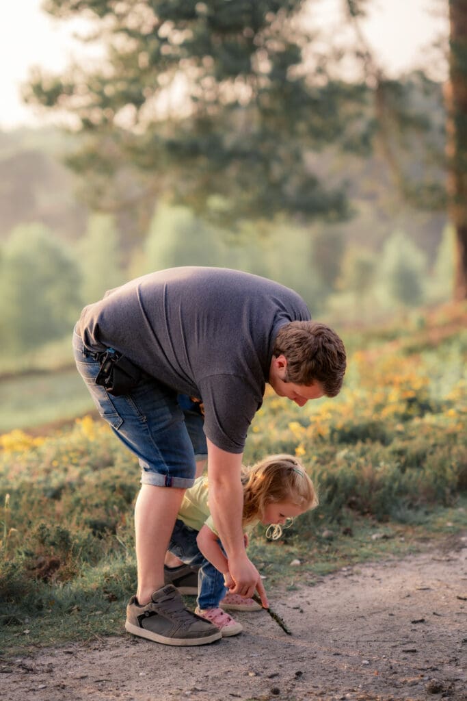 A dad and his tiny toddler both bent over examining something on the ground together on a sun-dappled heathland path