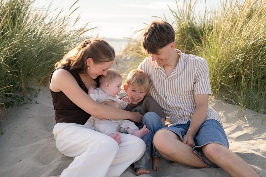 Mum holding baby close while toddler cuddles into dad, candid family moment in the dunes at West Wittering Beach.