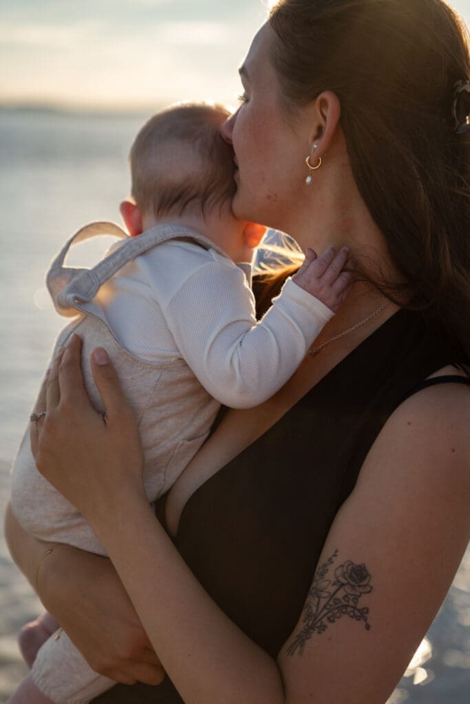 Mum kissing baby's head while standing at the water's edge during a family photoshoot in Sussex.