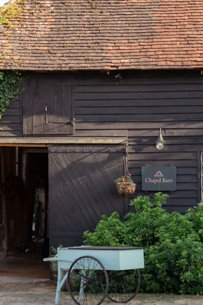 The black timber exterior and Chapel Barn sign with a vintage cart and hanging basket at the Bolney wedding venue in Sussex
