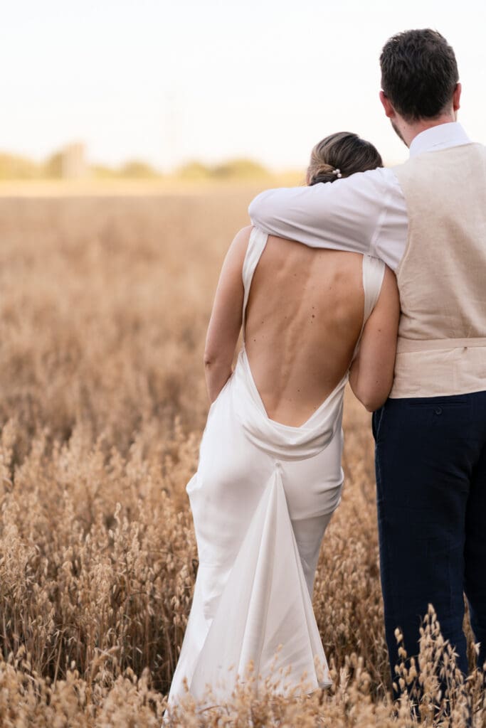 Bride in a backless satin gown with her groom's arm around her, looking out over a golden wheat field at sunset during their Chapel Barn wedding
