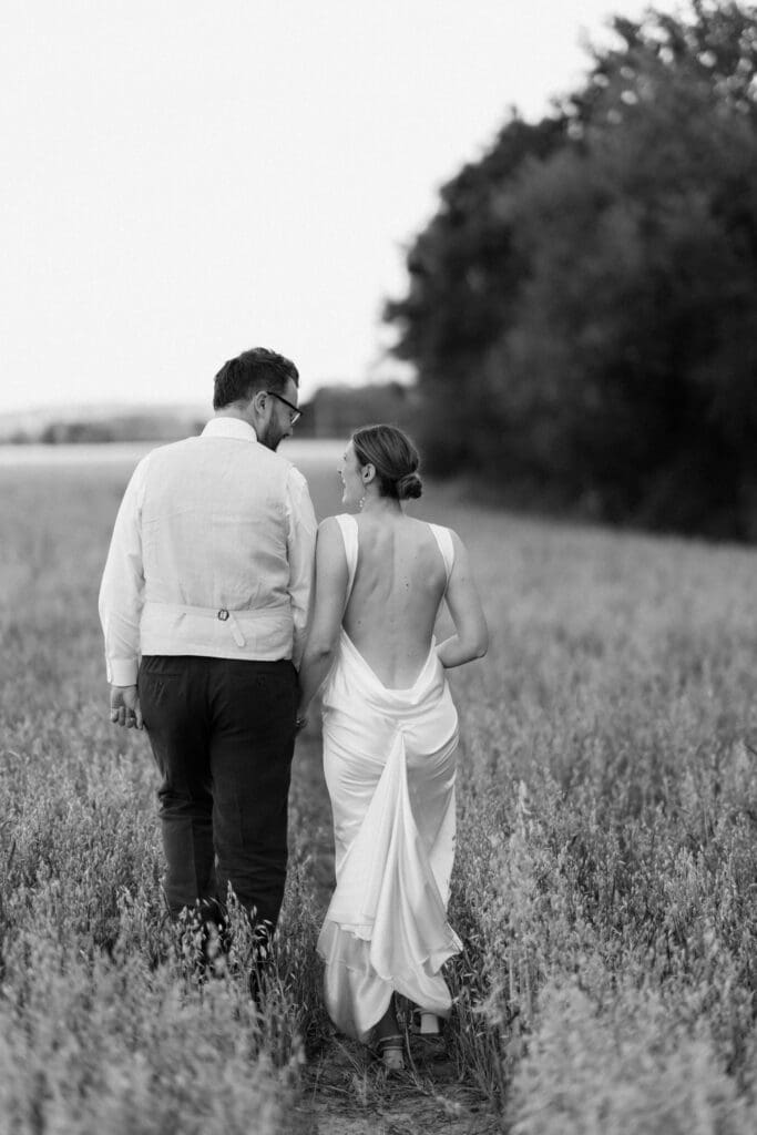 Black and white photo of the bride and groom walking hand in hand through a wheat field at sunset, Chapel Barn, Sussex
