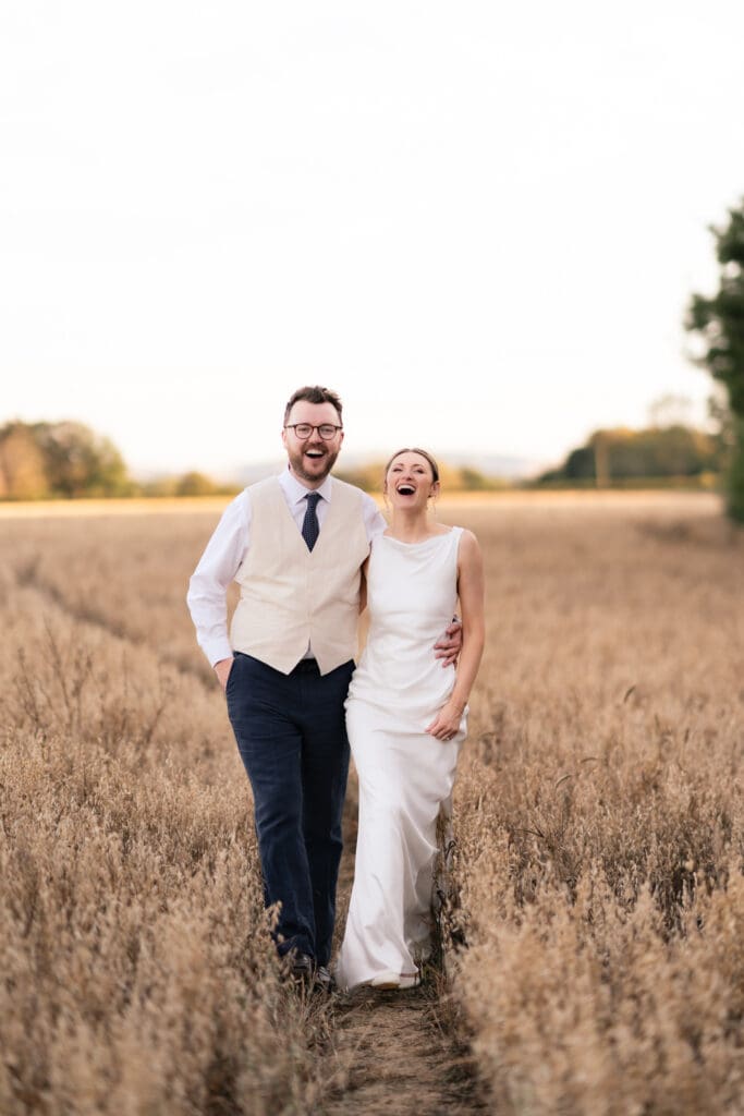 Bride and groom laughing as they walk together through a sunlit wheat field during their Chapel Barn wedding in Sussex