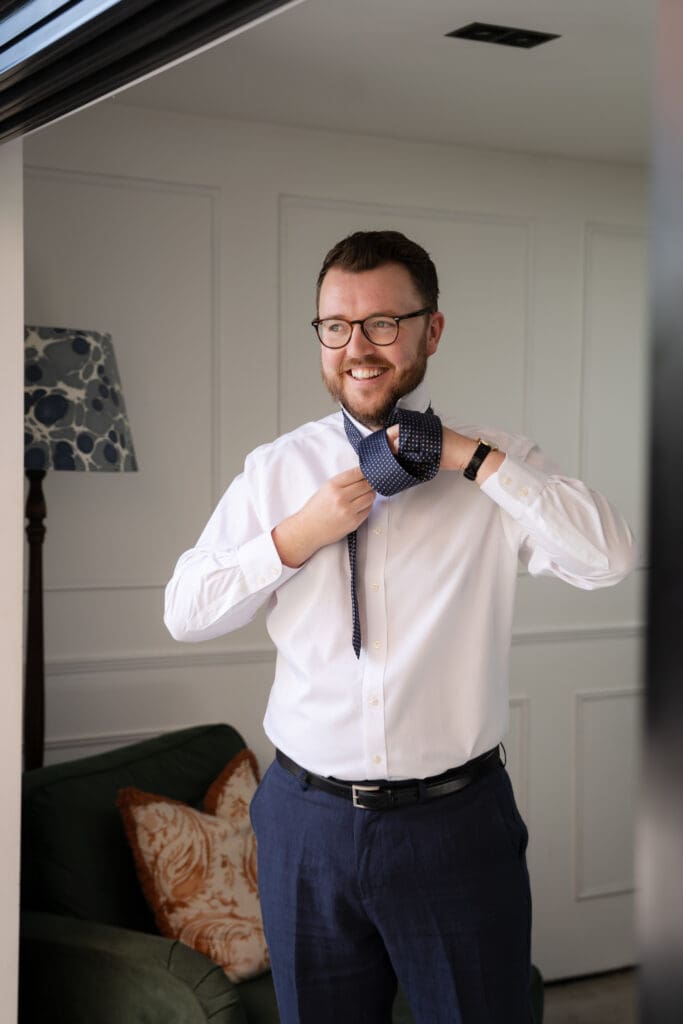Groom fastening his tie and smiling as he gets ready on the morning of his Chapel Barn wedding