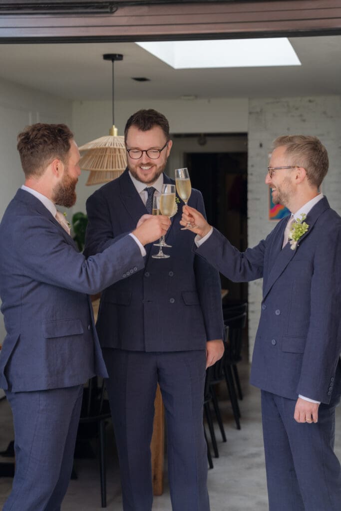 Groom and his two groomsmen toasting with champagne in navy suits while getting ready before the wedding