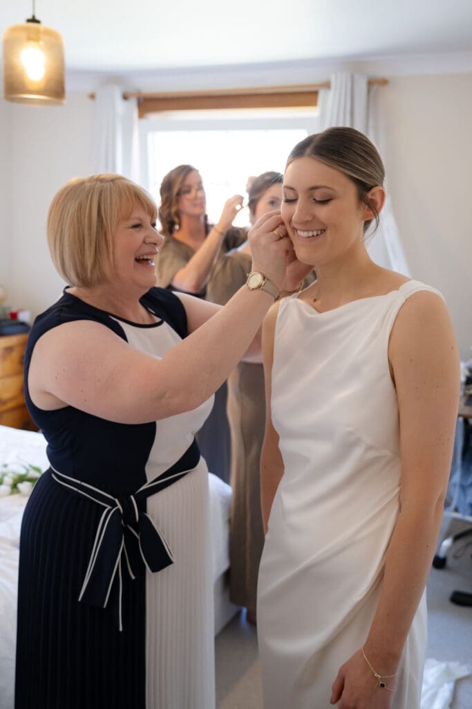 Bride laughing as her mum helps with her earrings while her bridesmaids get ready in the background on the wedding morning