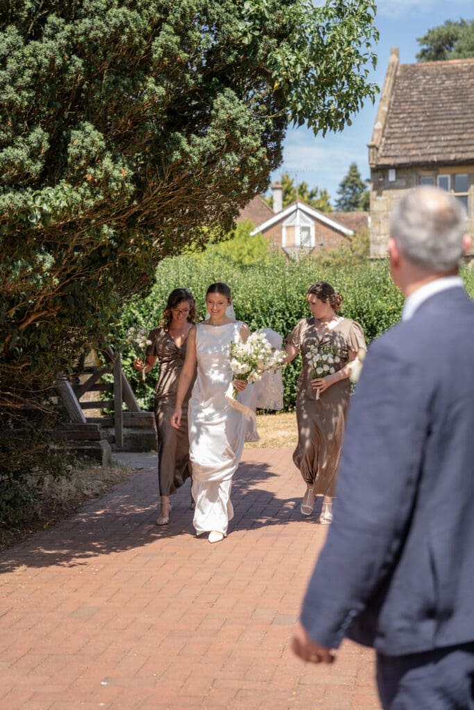 Bride walking to the church with her two bridesmaids in taupe dresses carrying white bouquets, at Holy Trinity Church, Cuckfield