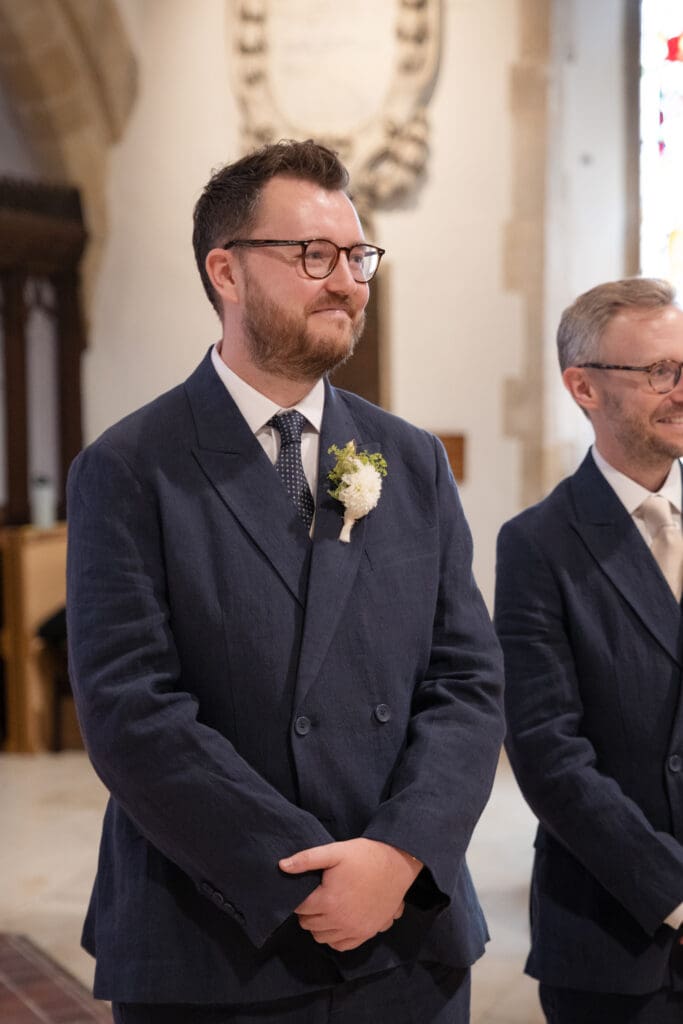 Groom smiling as he waits with his best man at the front of Holy Trinity Church, Cuckfield, during the ceremony