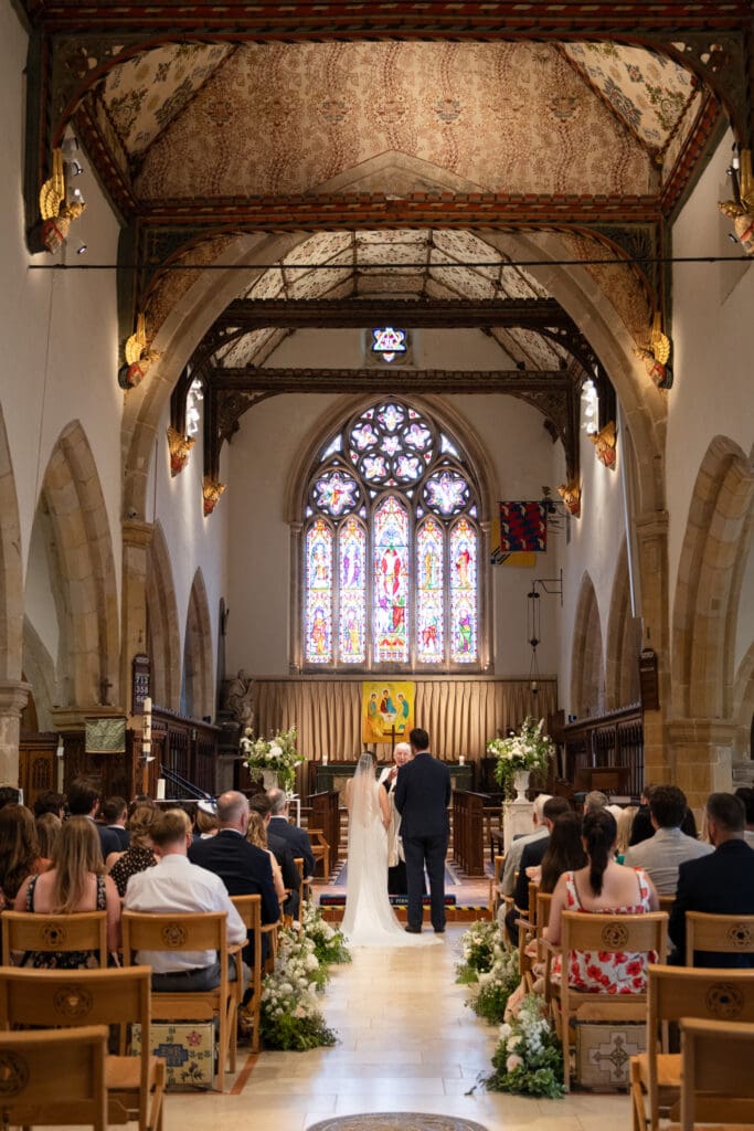 Bride and groom at the altar during their wedding ceremony inside Holy Trinity Church, Cuckfield, with stained glass window and floral aisle displays