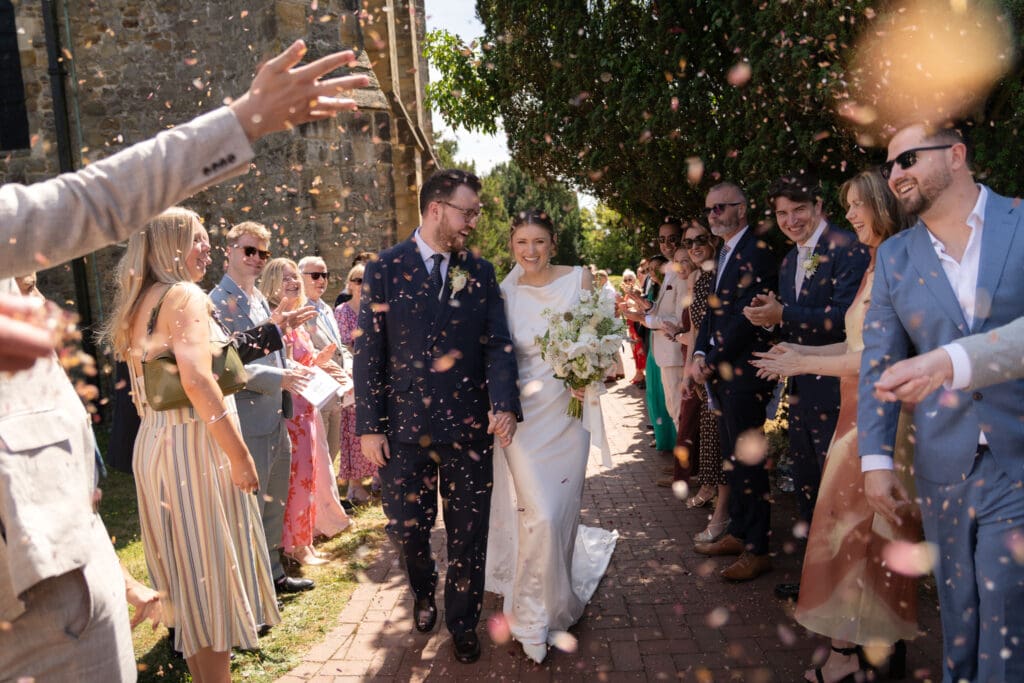 Bride and groom walking through a confetti throw as guests celebrate outside Holy Trinity Church, Cuckfield, Sussex