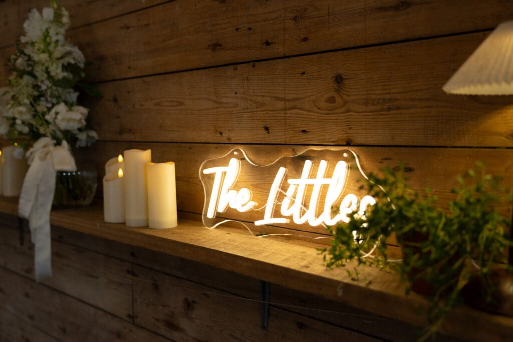 Personalised 'The Littles' neon sign glowing on a wooden shelf beside candles and white flowers inside Chapel Barn, Bolney