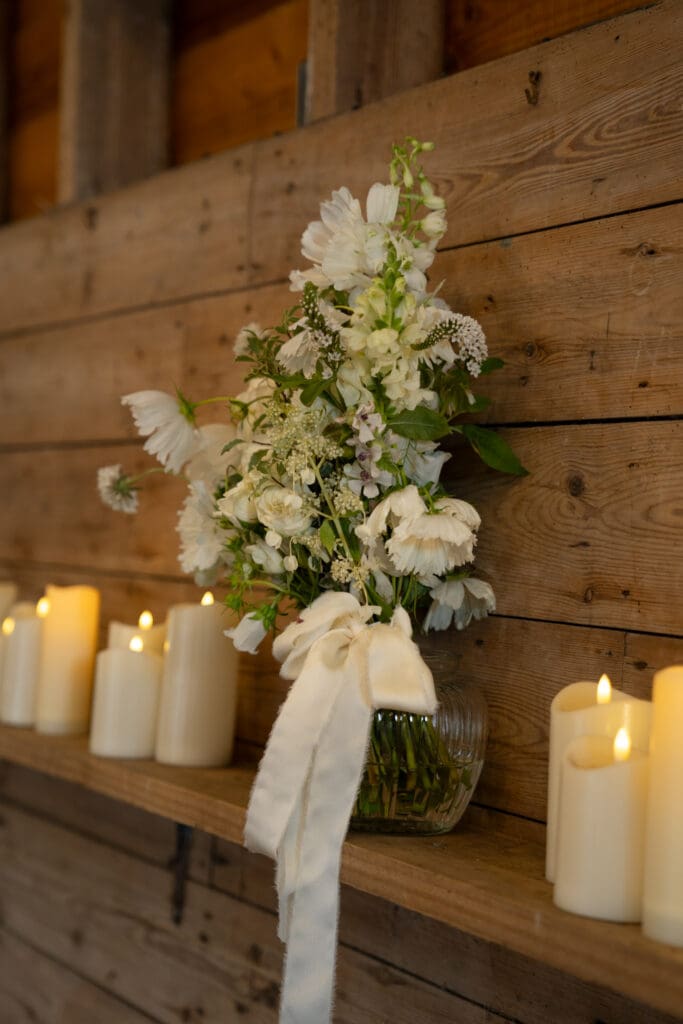 White and cream wedding flowers tied with silk ribbon on a wooden shelf beside candles inside Chapel Barn, Bolney
