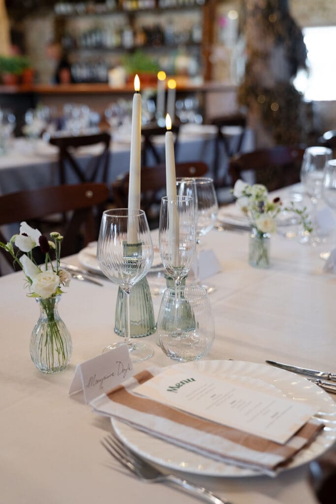 Wedding table setting with white roses in a bud vase, taper candles, ribbed glassware and a handwritten place name at Chapel Barn, Sussex