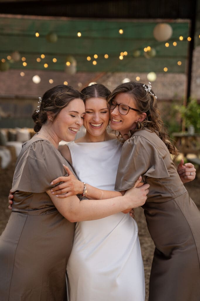 Bride embracing her two bridesmaids in taupe dresses under festoon lights during the evening reception at Chapel Barn, Sussex