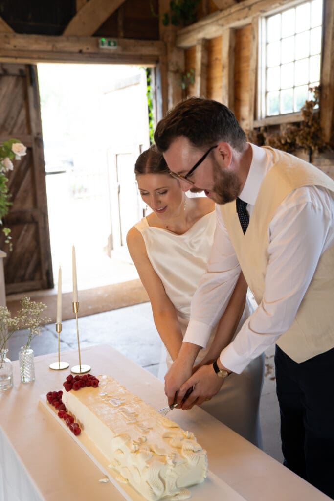 Bride and groom cutting their raspberry-topped wedding cake together inside Chapel Barn, Sussex