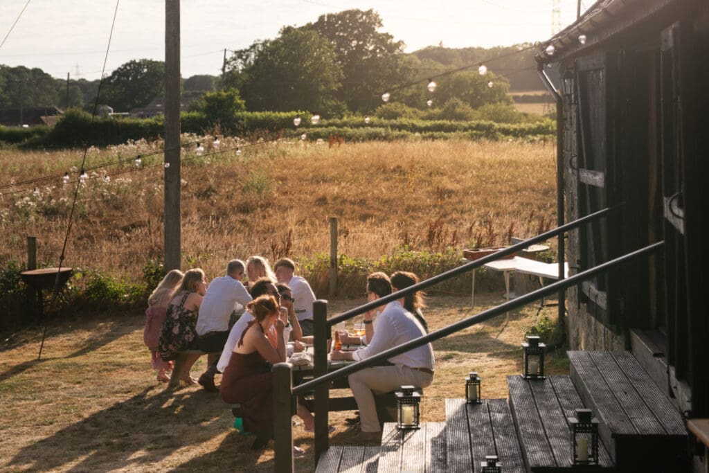 Wedding guests gathered around an outdoor table in golden evening light beside the barn and open Sussex countryside at Chapel Barn