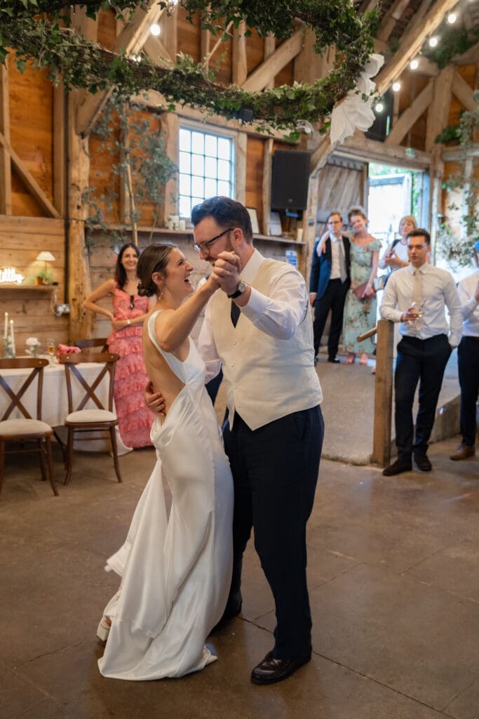 Bride and groom sharing their first dance beneath the foliage hoop as guests look on inside Chapel Barn, Bolney