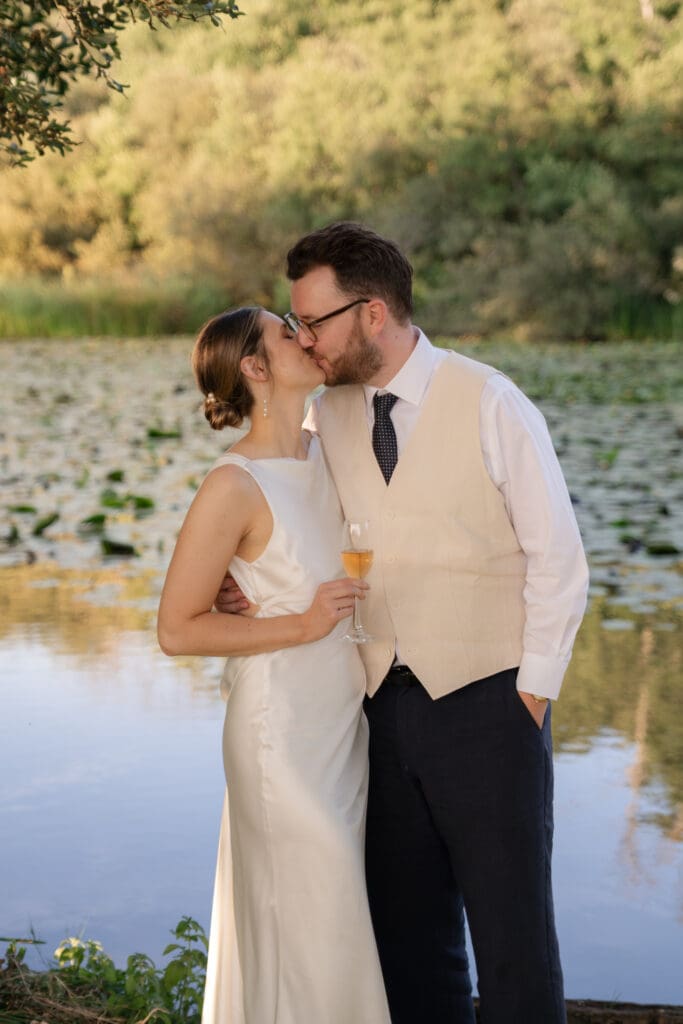 Bride and groom sharing a kiss beside the lily-covered lake at golden hour, the bride holding a glass of fizz, during their Chapel Barn wedding in Bolney, Sussex