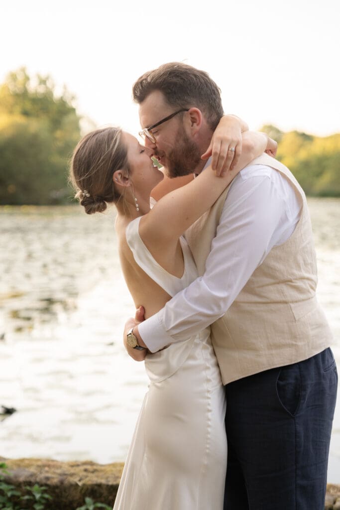 Bride and groom sharing a kiss with arms wrapped around each other beside the lily lake at golden hour, Chapel Barn, Sussex