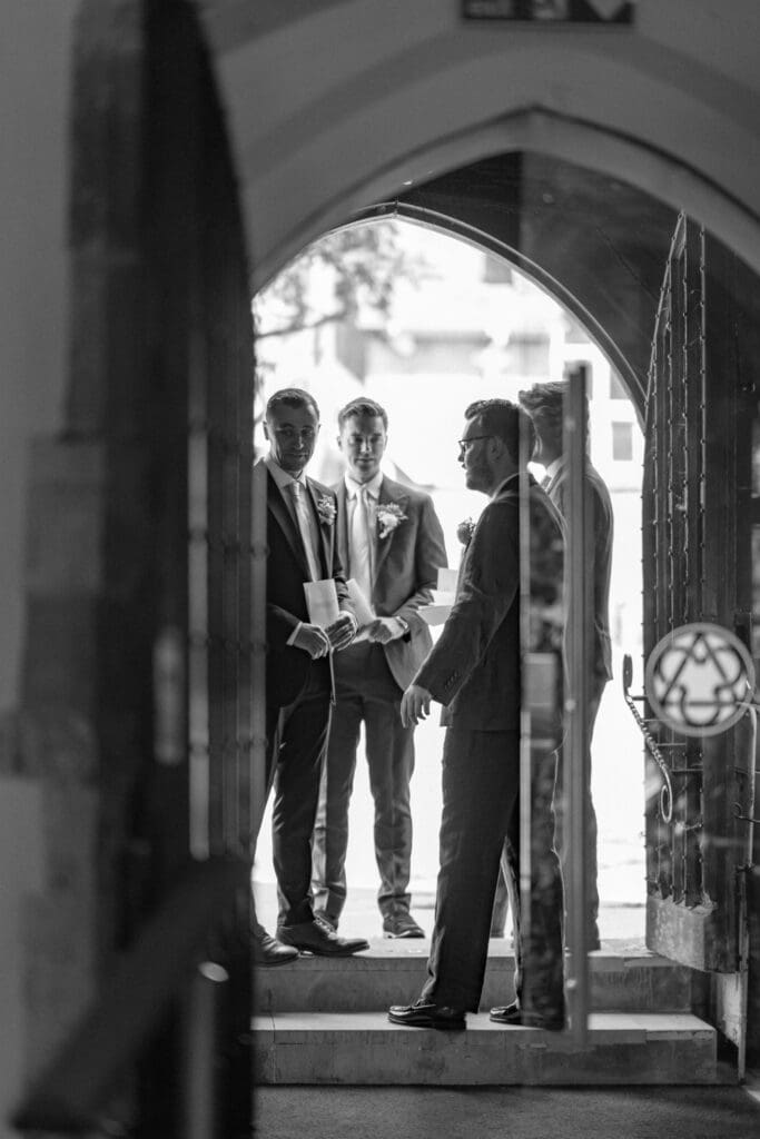 Black and white photo of the groom and groomsmen waiting in the doorway of Holy Trinity Church, Cuckfield
