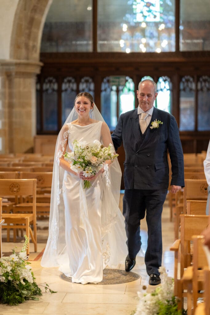Bride walking down the aisle with her father, holding a wild white bouquet, at Holy Trinity Church, Cuckfield, Sussex