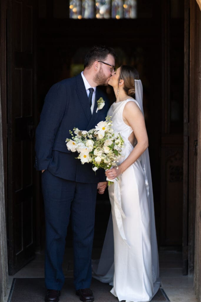 Bride and groom kissing in the doorway of Holy Trinity Church, the bride holding her wild white bouquet, Cuckfield, Sussex