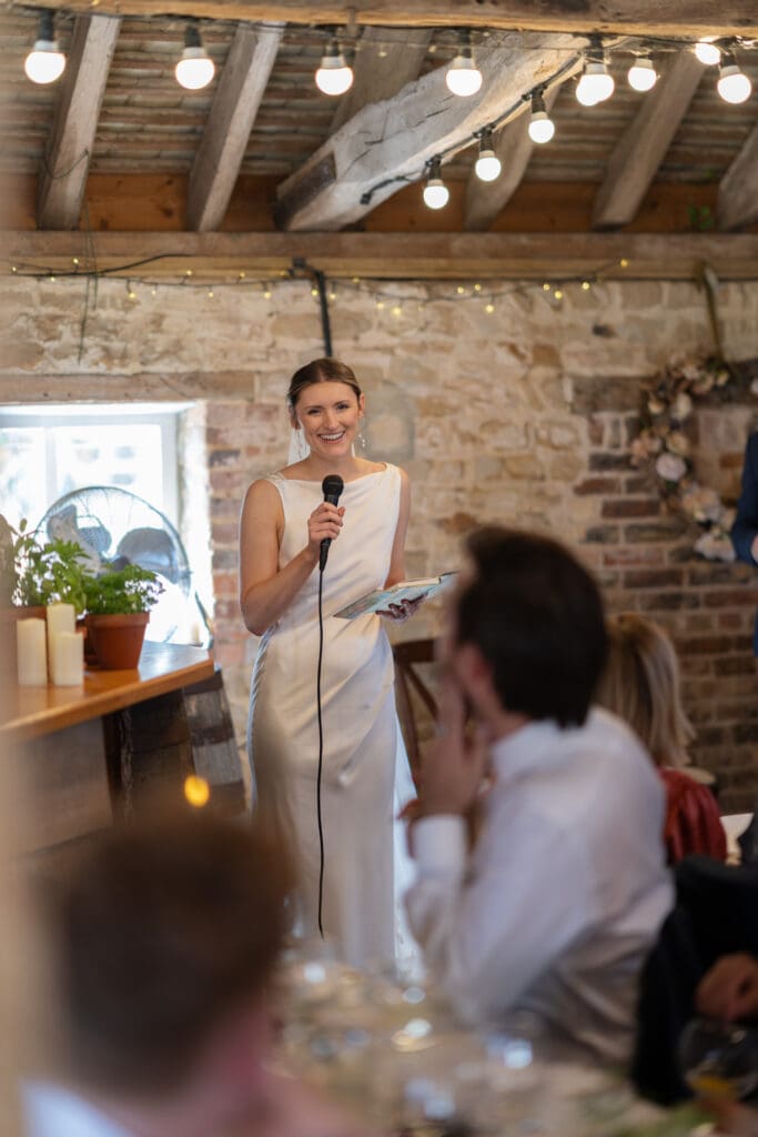 Bride laughing as she gives her speech into a microphone beneath the oak beams and festoon lights inside Chapel Barn, Bolney